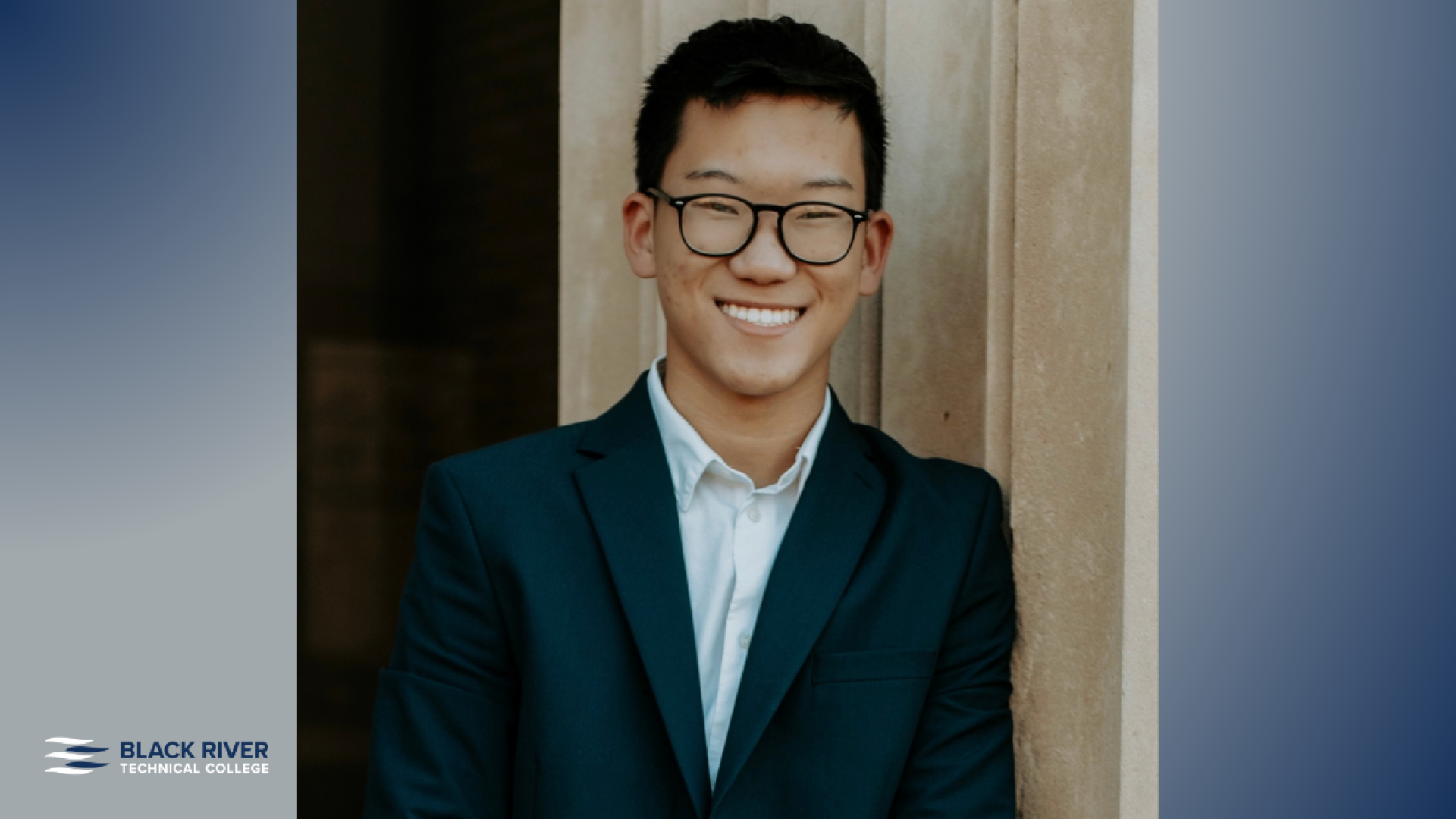 A young man with short dark hair and glasses, wearing a dark suit jacket and white shirt, smiles while leaning against a stone column, with Black River Technical College branding.